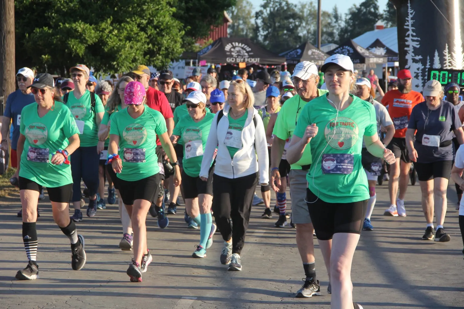 A group of runners, mostly women in green shirts, running on a paved road with tents and a finish line arch in the background.