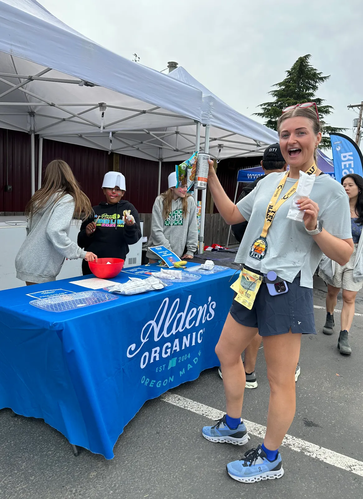 A woman wearing a race medal and holding snacks smiles excitedly at the camera, standing in front of a table with volunteers.