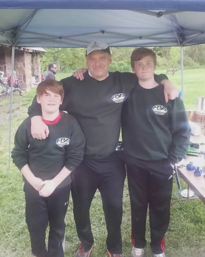 Three people, two boys and an adult male, posing for a photo under a tent at an outdoor event.