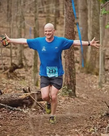 A bald runner with arms outstretched runs on a wooded trail, wearing a blue shirt with bib number 253.