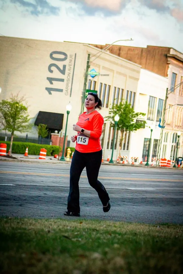 A female runner wearing an orange long-sleeved shirt and black pants runs on a city street, with a bib number 115 visible.