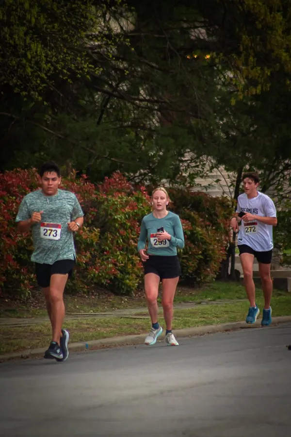 Three runners on a road course, with trees and bushes in the background.