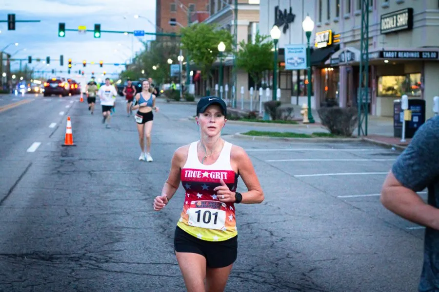 A female runner wearing a white tank top with red, white, and blue stars and the words 'TRUE GRIT' runs down a city street during a race. She has the number 101 on her bib.