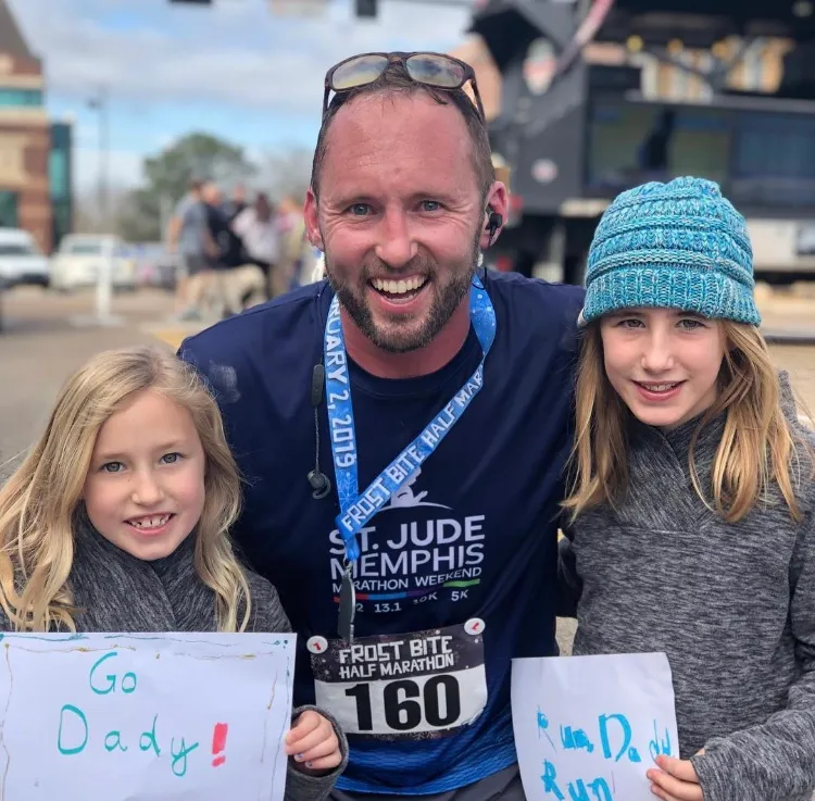 A smiling man wearing a race bib and medal stands with his two daughters, who are holding signs that say 'Go Daddy!' and 'Run Daddy Run'.