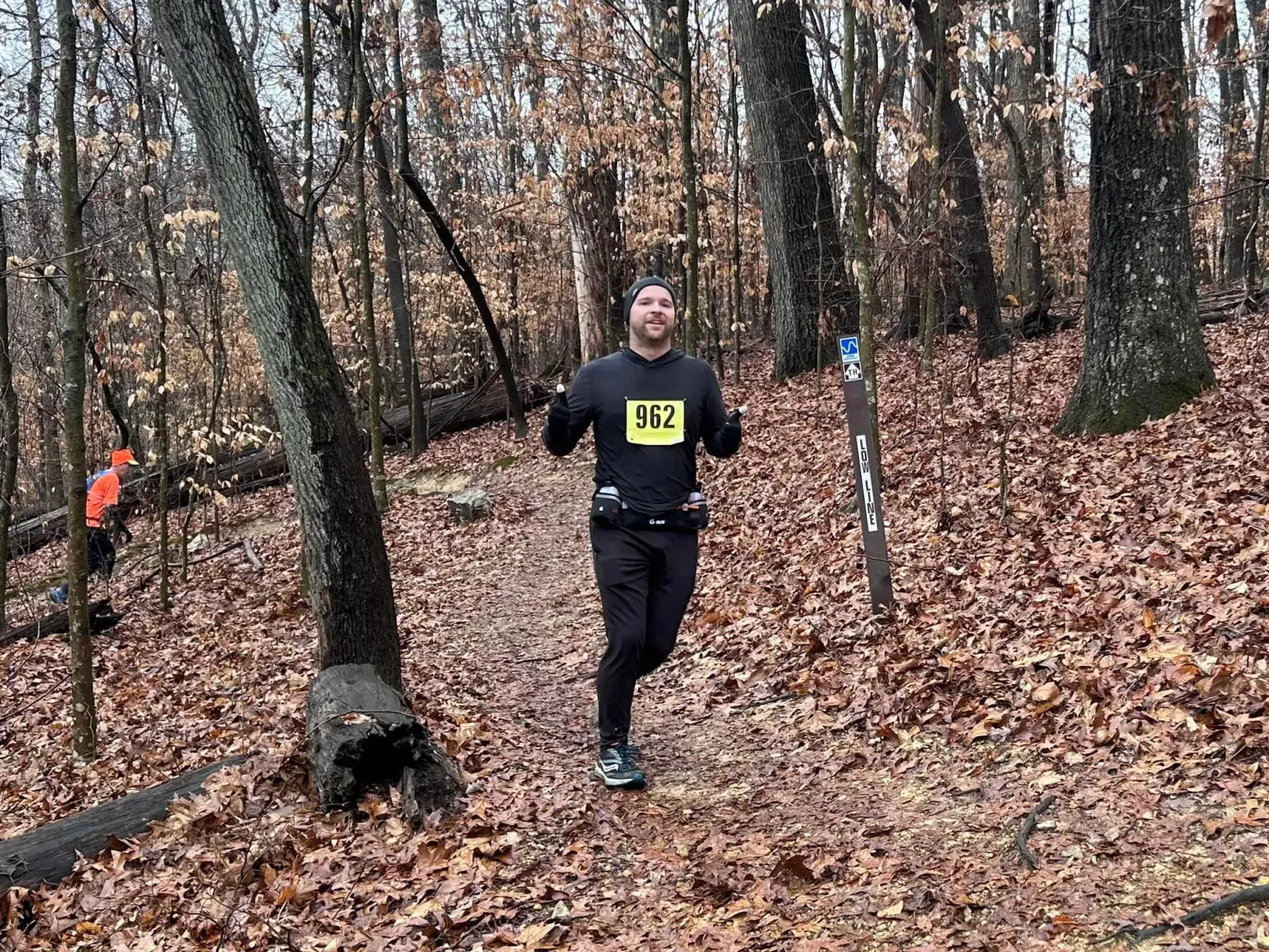 A male runner with bib number 962 smiles and gives a thumbs up while running on a leaf-covered trail in a forest.
