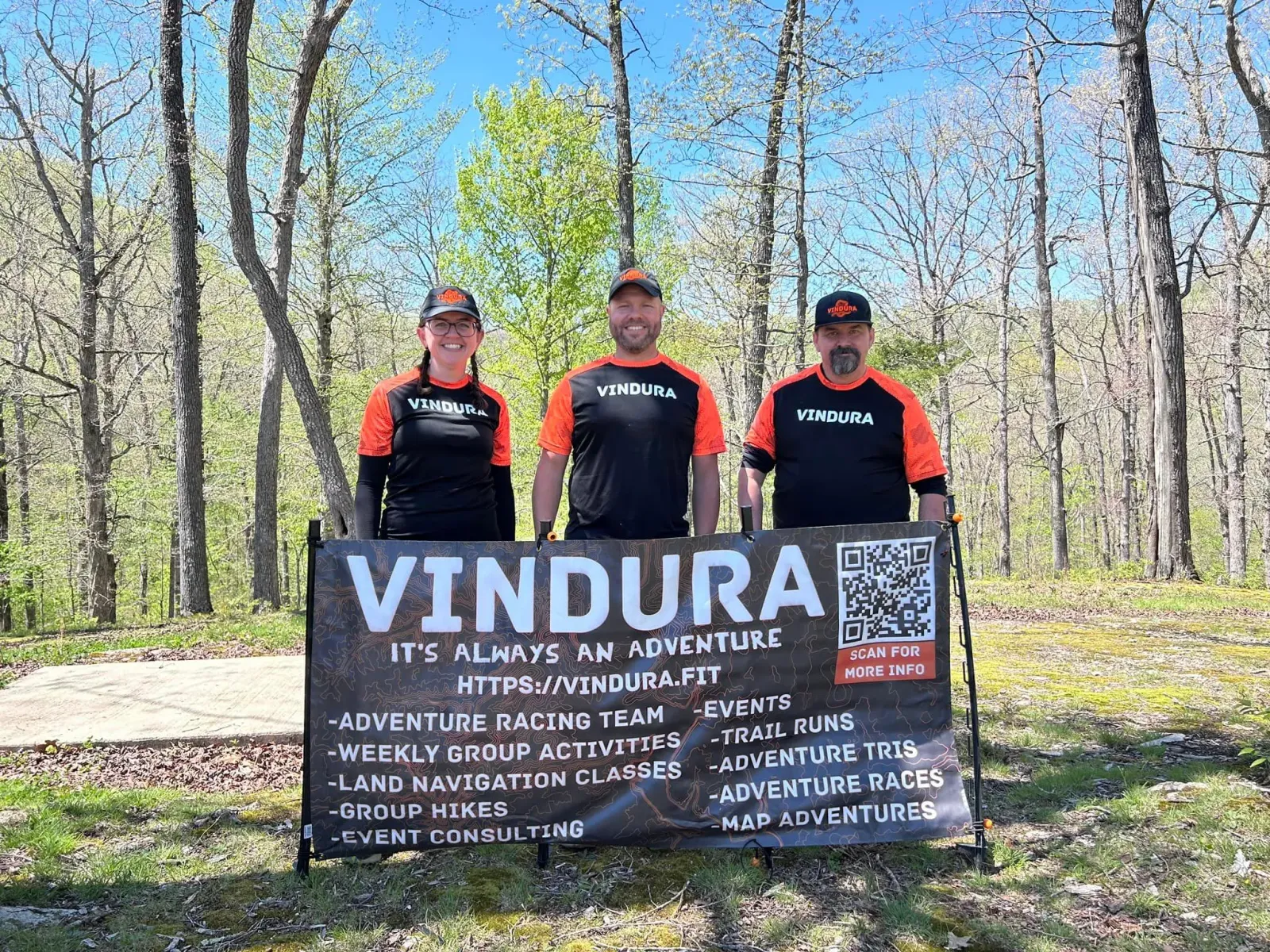 Three people wearing Vindura branded shirts and hats stand behind a large banner that advertises Vindura's services, including trail runs and adventure races.