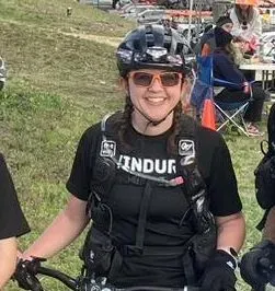 A smiling woman wearing a helmet and sunglasses, dressed in athletic gear, poses with her bicycle.