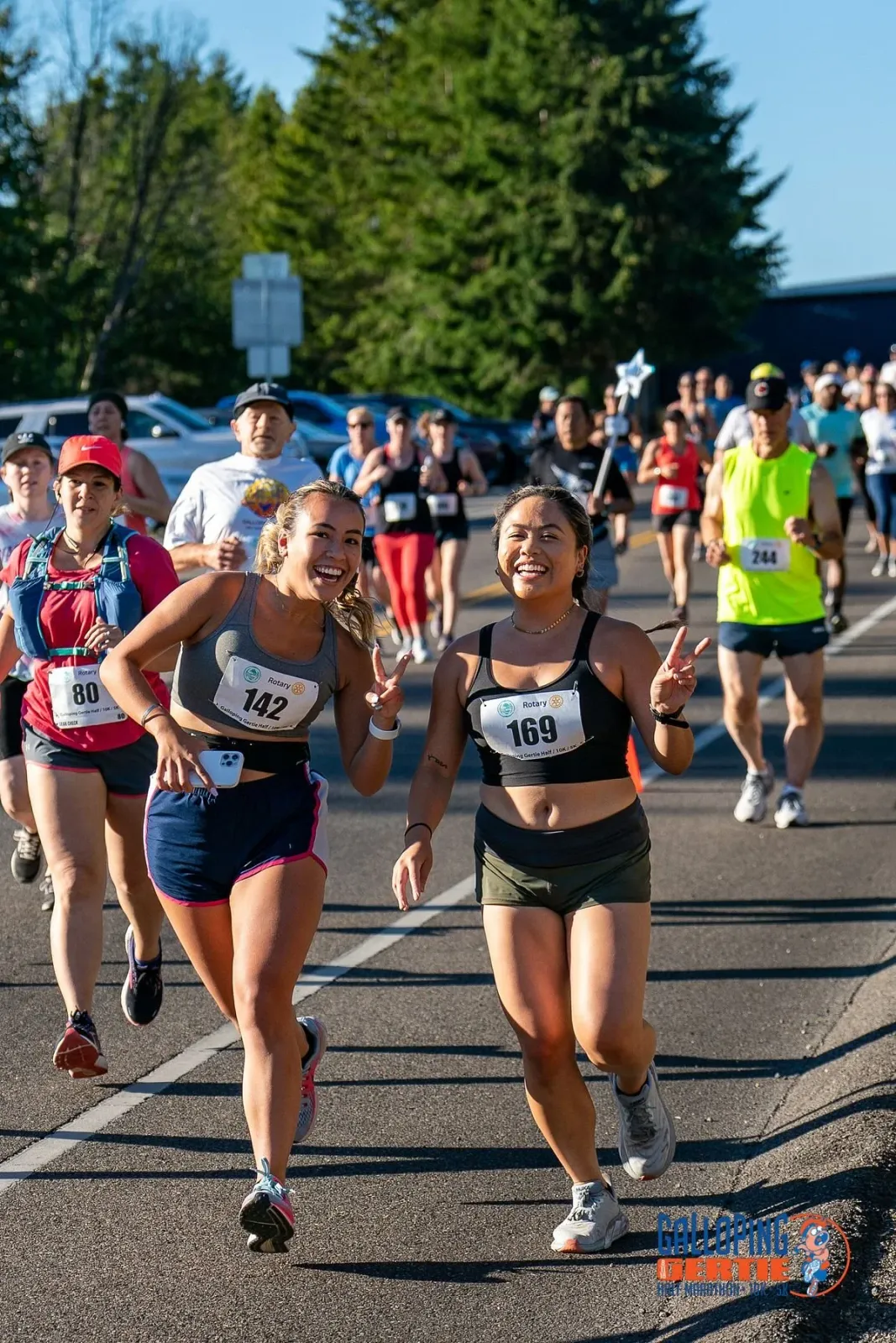 Two female runners smiling and giving peace signs while running in a race, with other participants in the background.