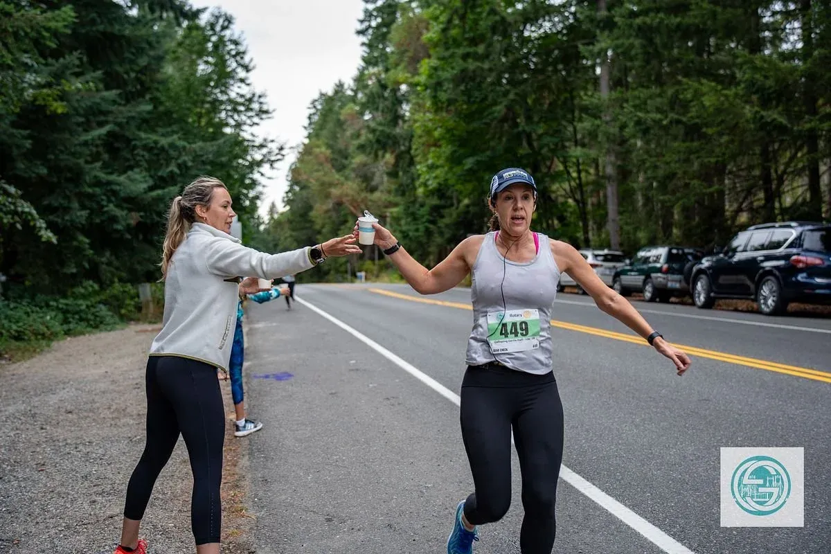 A female runner with bib number 449 receives a cup from a volunteer on the side of a tree-lined road during a race.