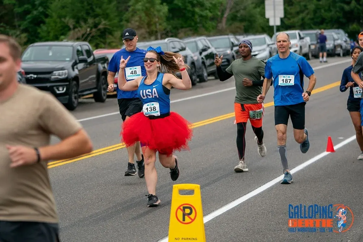 A female runner in a red tutu and USA-themed top smiles and makes a peace sign while running, with other runners and parked cars in the background.