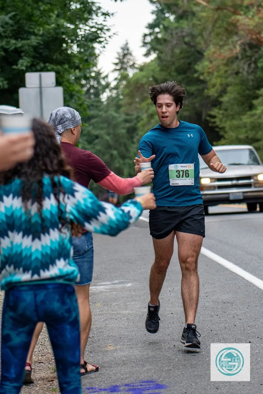 A male runner with bib number 376 is taking a cup from a volunteer on a tree-lined road during the Galloping Gertie Half Marathon.