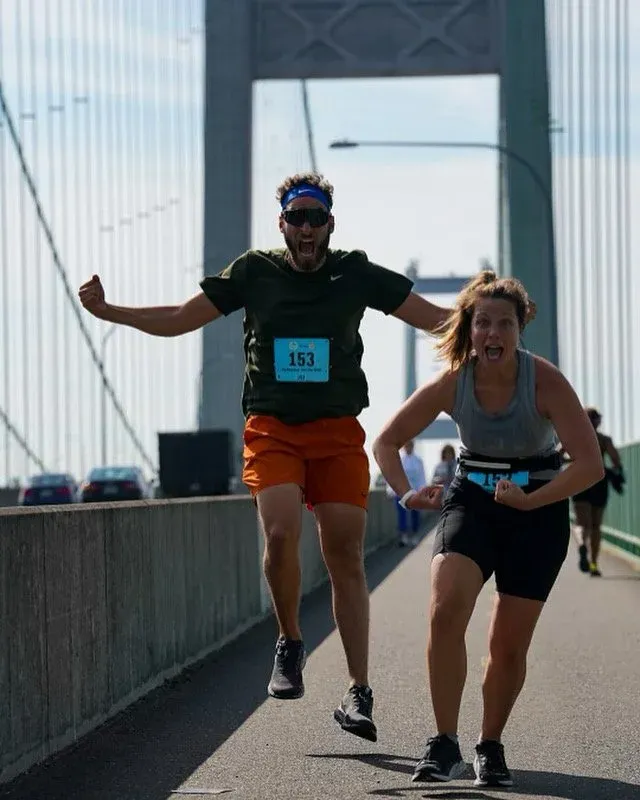 Two runners, one male and one female, are joyfully running and celebrating on a bridge during a race. The male runner is mid-air with his arms outstretched, and the female runner is flexing her bicep. They are both wearing race bibs.