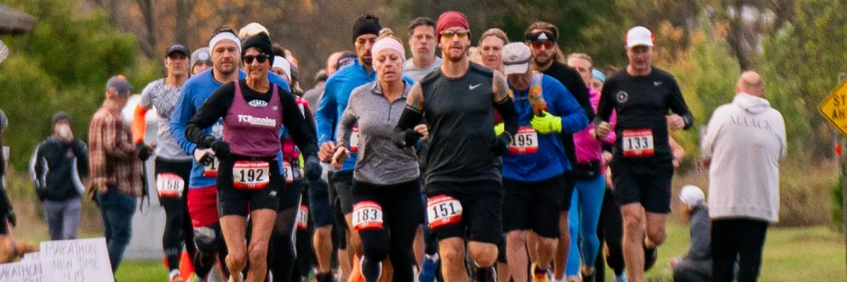 A group of runners in athletic wear, some with bib numbers, are running forward at the start of a race.