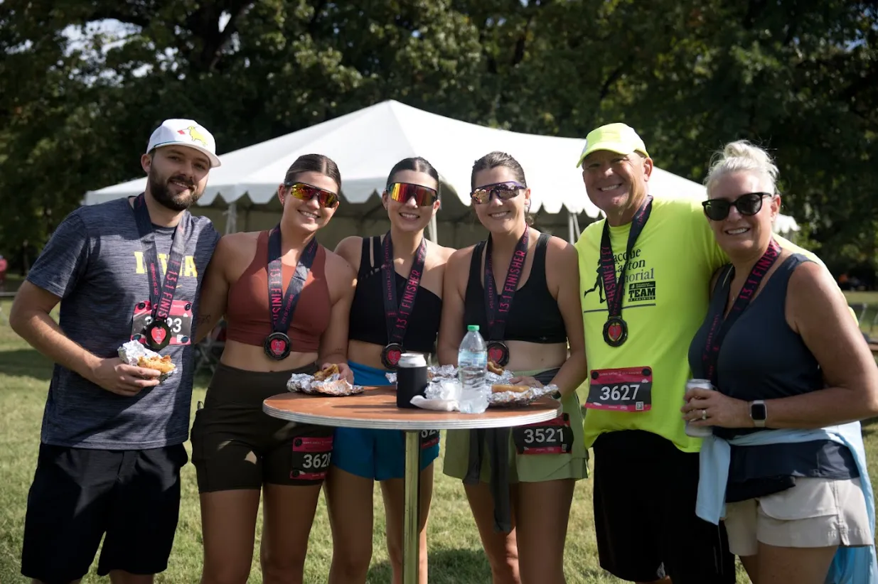 A group of six people, some wearing race medals and bibs, smiling and holding food and drinks after completing a half marathon.