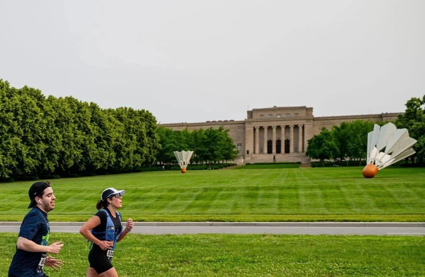 Two runners, a man and a woman, are running on a road with a large building and sculptures in the background.