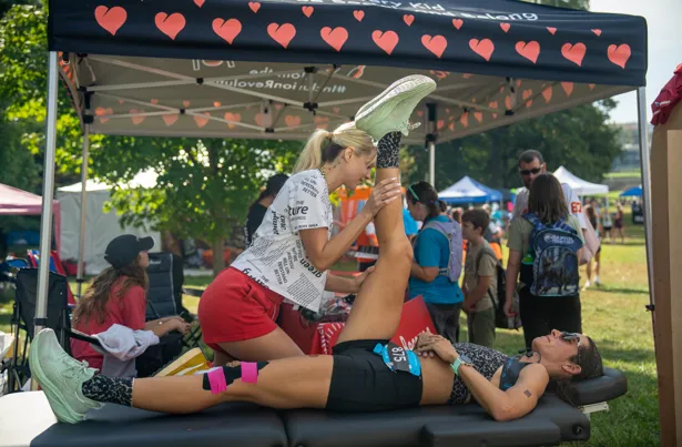 A runner lies on a massage table while a therapist stretches her leg, with other people and tents in the background.