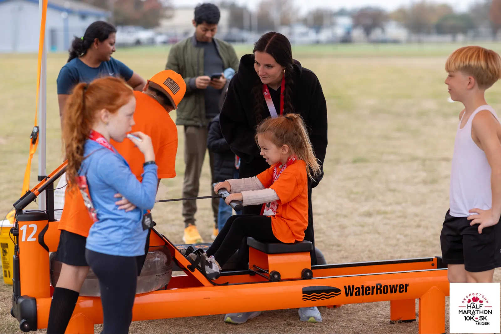 Children and adults gathered around a rowing machine at a race event, with one child actively using it.