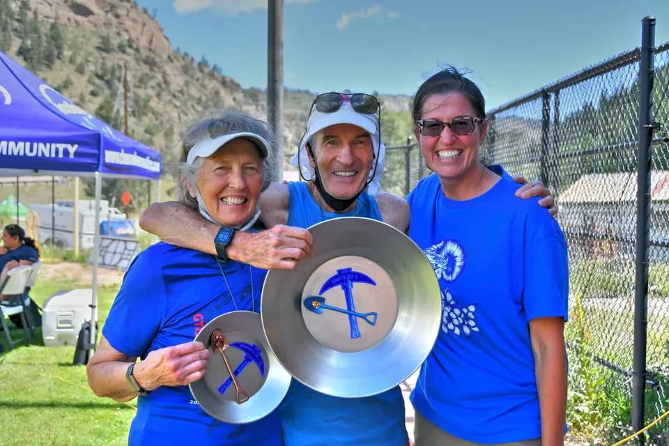 Three smiling runners, two women and one man, pose for a photo holding large gold pans with mining tools inside, representing awards.