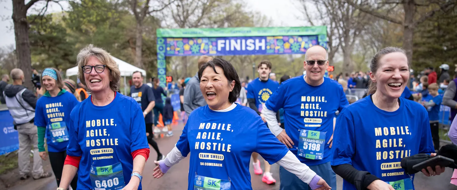 A group of smiling runners in blue t-shirts with 'MOBILE, AGILE, HOSTILE' printed on them, celebrating at the finish line of a race.
