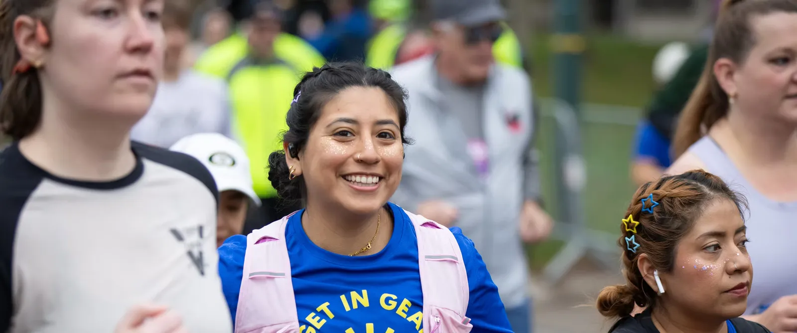Runners smiling and participating in a race, with one runner in the foreground wearing a blue shirt that says "GET IN GEAR".