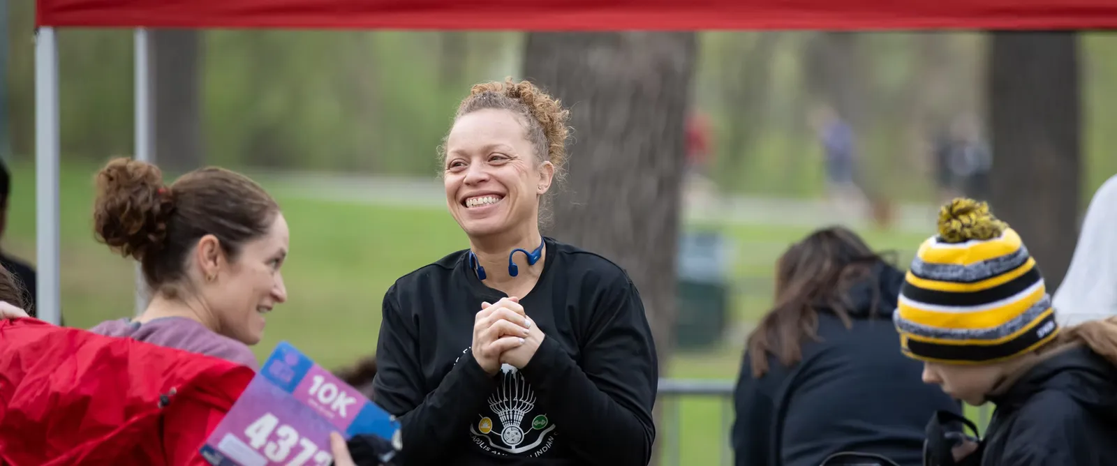 A woman with curly hair smiles broadly while looking to her left, with other participants blurred in the background at a race event.