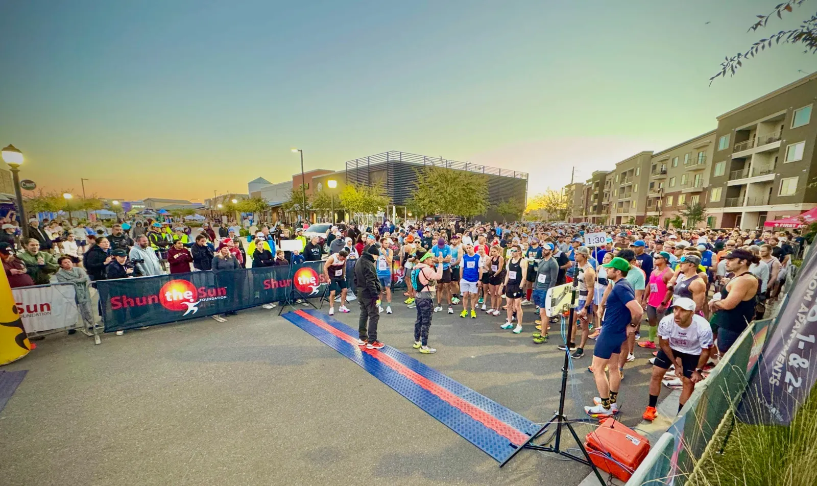 A large group of runners gathered at the start line of a race, with buildings and a clear sky in the background.