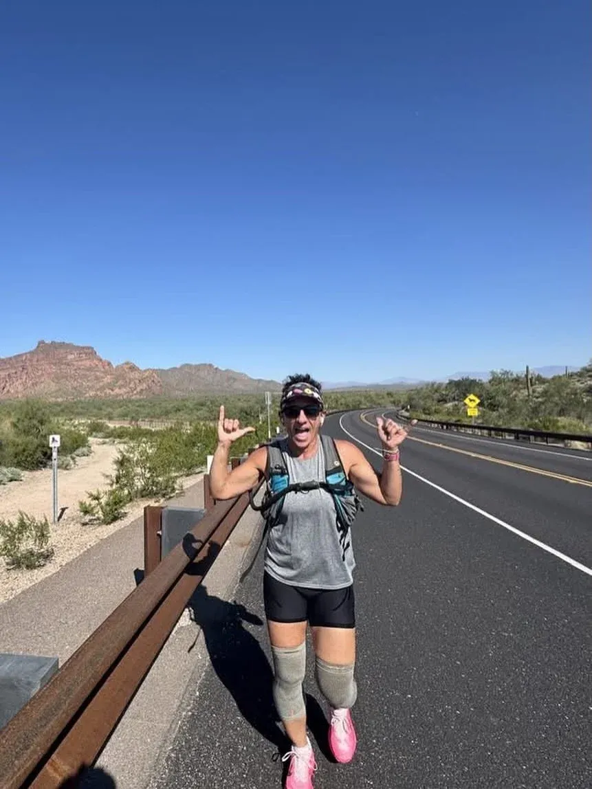 A runner smiles and makes 'rock on' hand gestures while running on a road with desert mountains in the background.