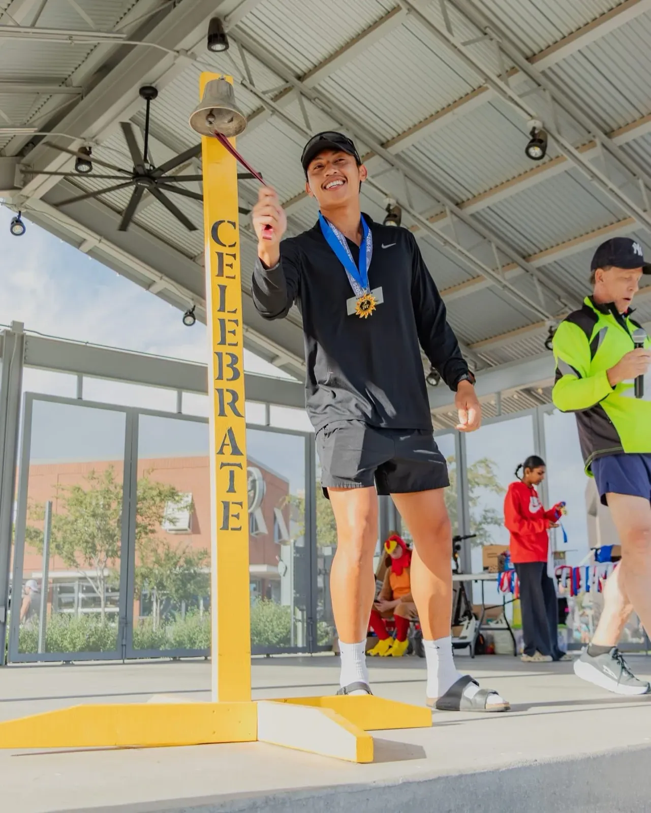 A smiling runner wearing a medal rings a bell on a yellow post labeled 'CELEBRATE'.