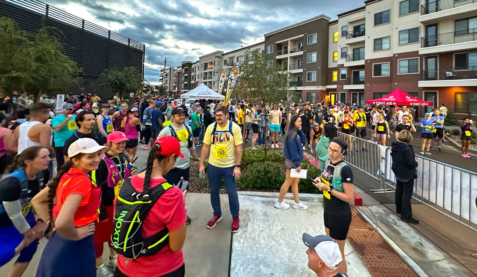 A large group of runners gathered before the start of a race, with apartment buildings in the background and tents set up for race services.
