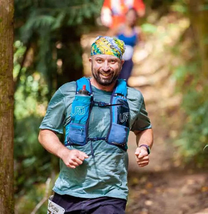 A male runner with a bandana smiles while running on a wooded trail during a race.
