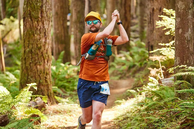 A male runner with a beard, wearing sunglasses and a yellow cap, runs on a forest trail, clapping his hands.