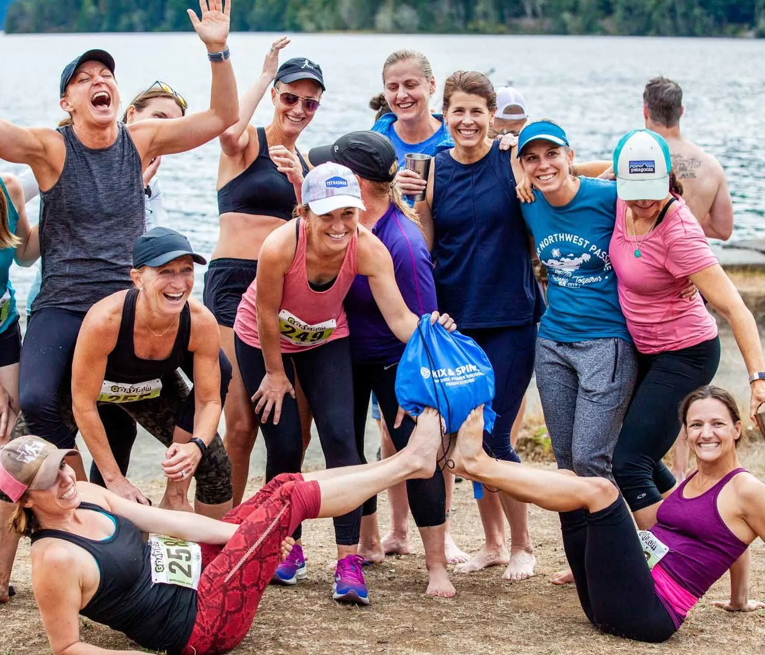 A group of smiling female runners celebrating by a lake, some are posing playfully.