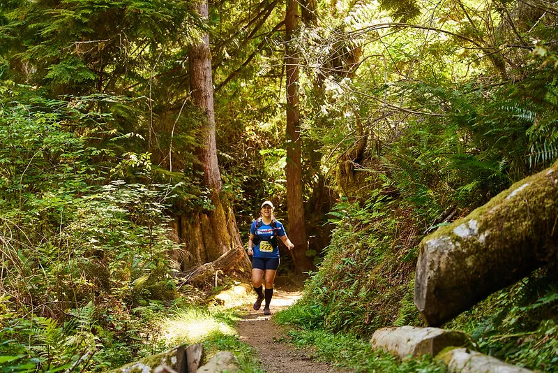 A female runner smiles as she runs on a dirt path through a lush, green forest.