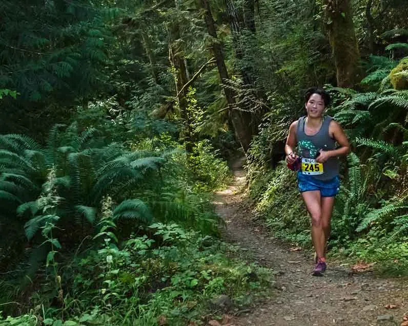 A female runner with bib number 245 runs on a dirt trail through a lush green forest.