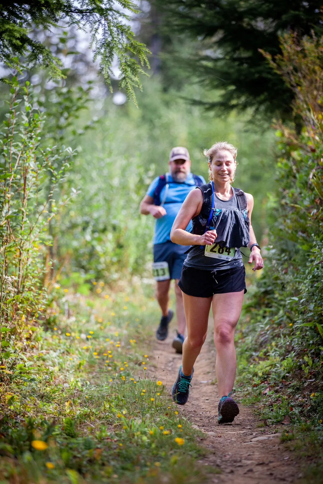 A female runner smiles as she runs on a dirt trail, with another runner behind her in a forest setting.