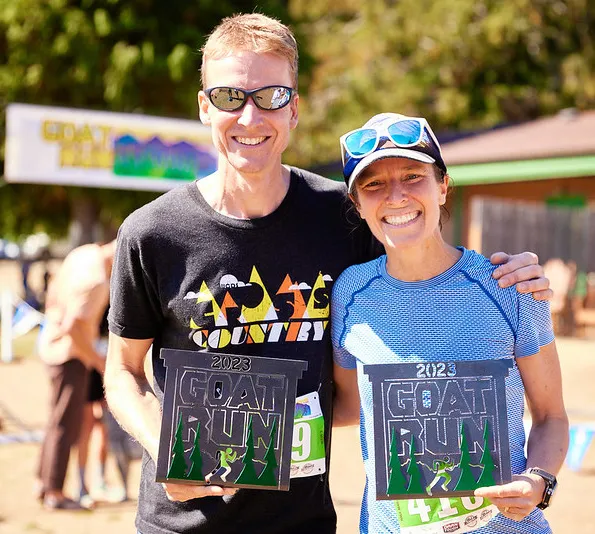 A smiling man and woman, both runners, pose for a photo holding awards from the Goat Run race.