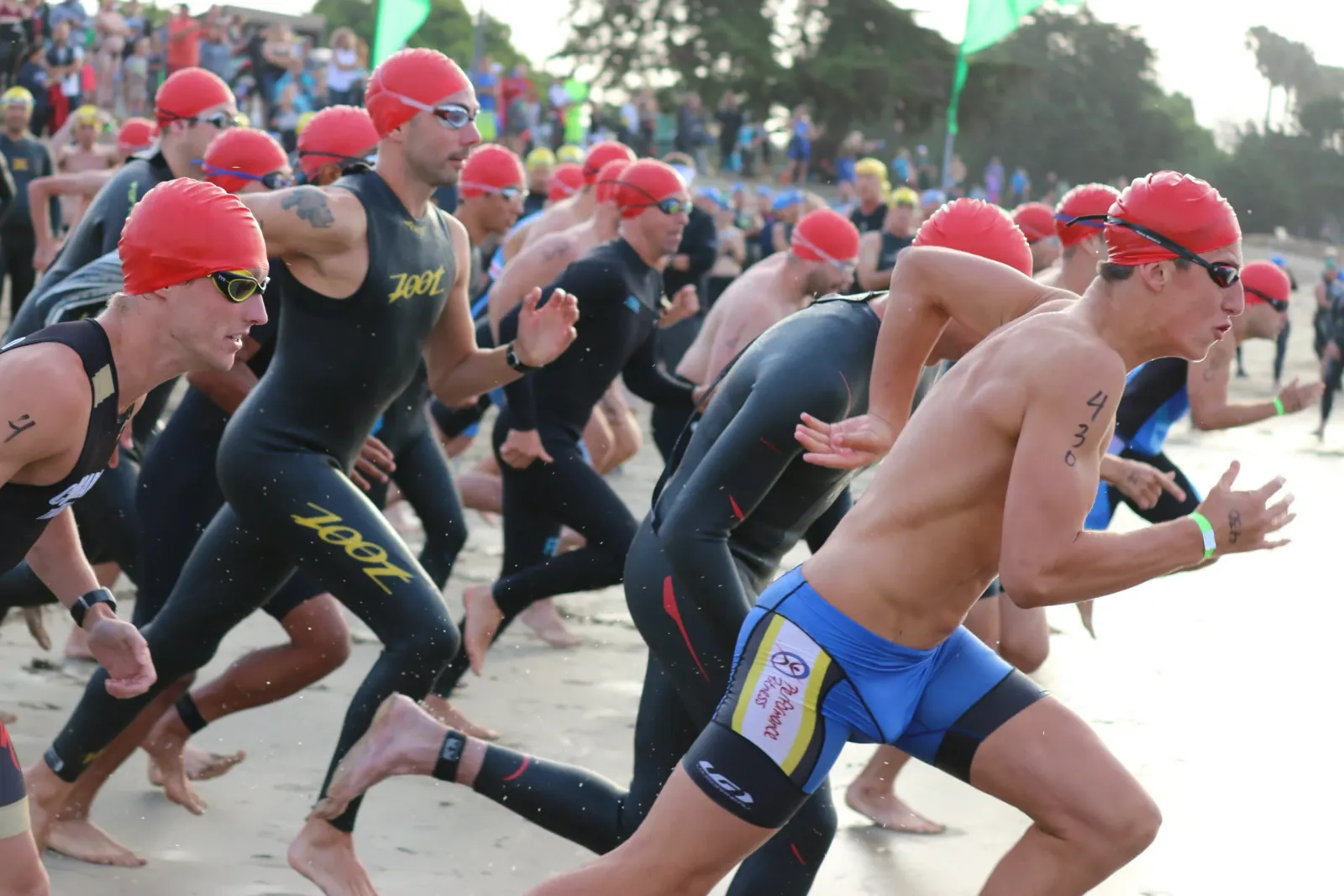 Runners in red caps and goggles starting a race on a sandy beach.
