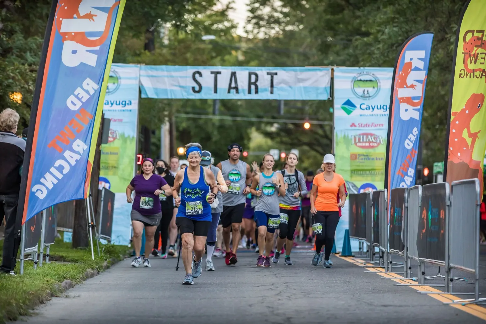 Runners crossing the start line at the Gorges Ithaca Half Marathon, with colorful banners and a 'START' sign overhead.