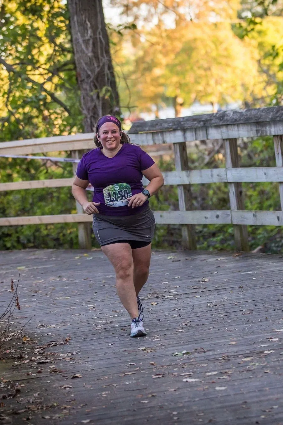 A female runner wearing a purple shirt and grey skirt smiles as she runs across a wooden bridge.