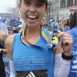 A smiling female runner proudly displays her race medal after finishing a road race.