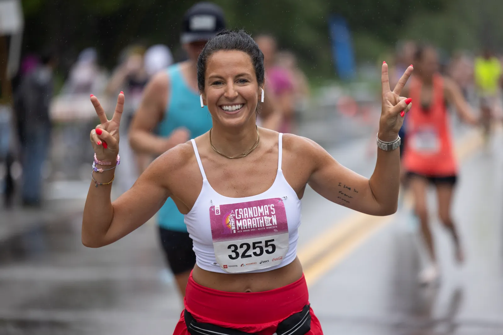 A female runner smiles and holds up two peace signs while running in Grandma's Marathon, with other runners blurred in the background.