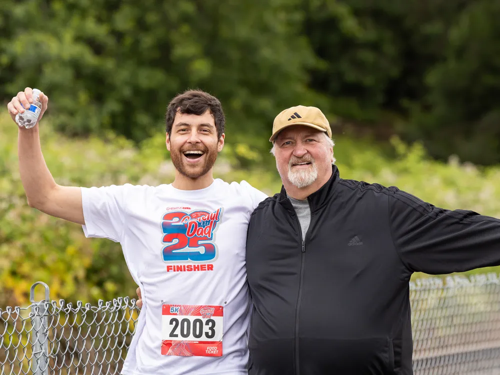 Two men smiling after completing a race, one holding a water bottle and wearing a 'Grateful Dad 25 FINISHER' shirt, the other wearing a baseball cap and a race bib.