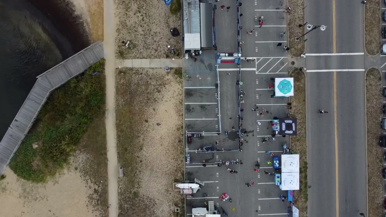 Aerial view of runners gathering at the start line of a road race, with tents and parking lot infrastructure visible.