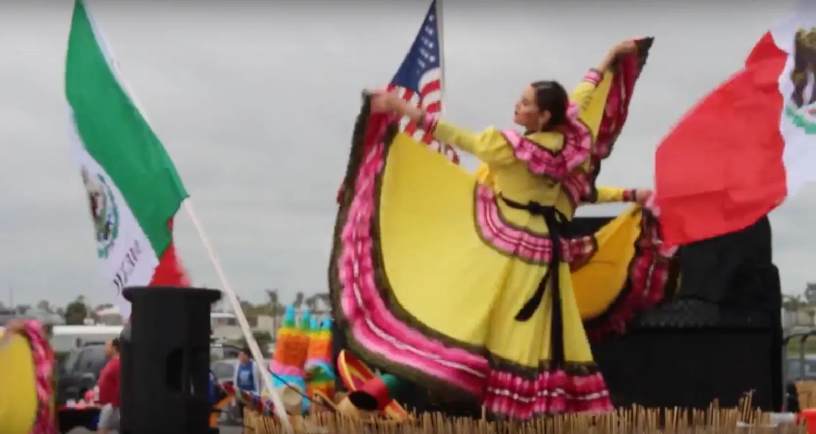 A woman in a yellow and pink traditional dress dances with flags, including the Mexican and American flags, in the background.