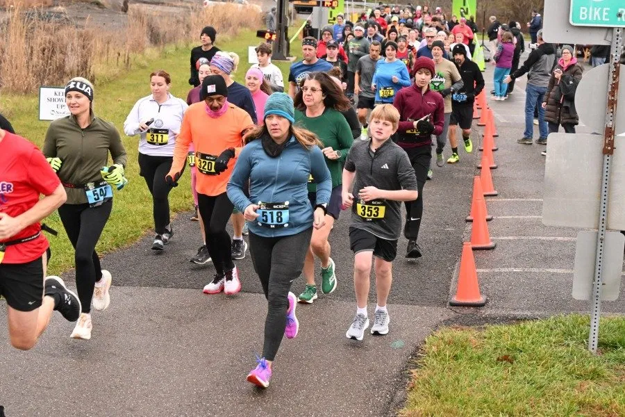 A group of runners, including men, women, and children, are running on a paved path lined with orange cones. Some runners are wearing bib numbers and athletic clothing.