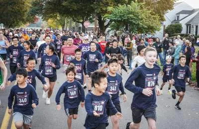 A group of young runners, mostly children, in matching dark blue shirts with race bibs, running down a street lined with trees and houses during a race.