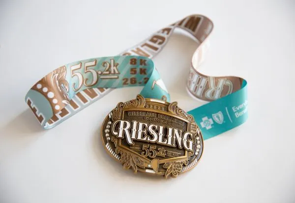 A close-up photo of a gold and bronze race medal with a teal and brown ribbon. The medal is oval-shaped and features the word "RIESLING" prominently in the center, along with text indicating "WINEGLASS CHALLENGE" and "55k". The ribbon has text and graphics, including "55k" and "26.2".