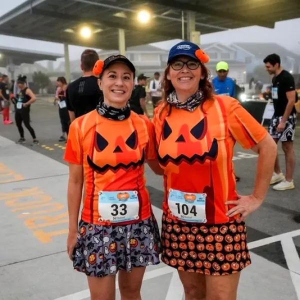 Two women in matching pumpkin-themed running outfits smile at the camera, with race bibs visible.