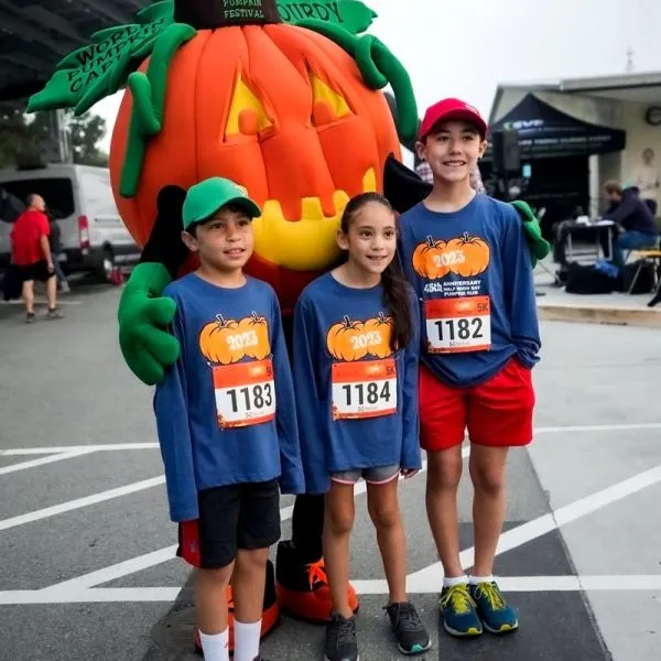 Three young runners in blue race t-shirts pose with a large pumpkin mascot at the Half Moon Bay Pumpkin Run.