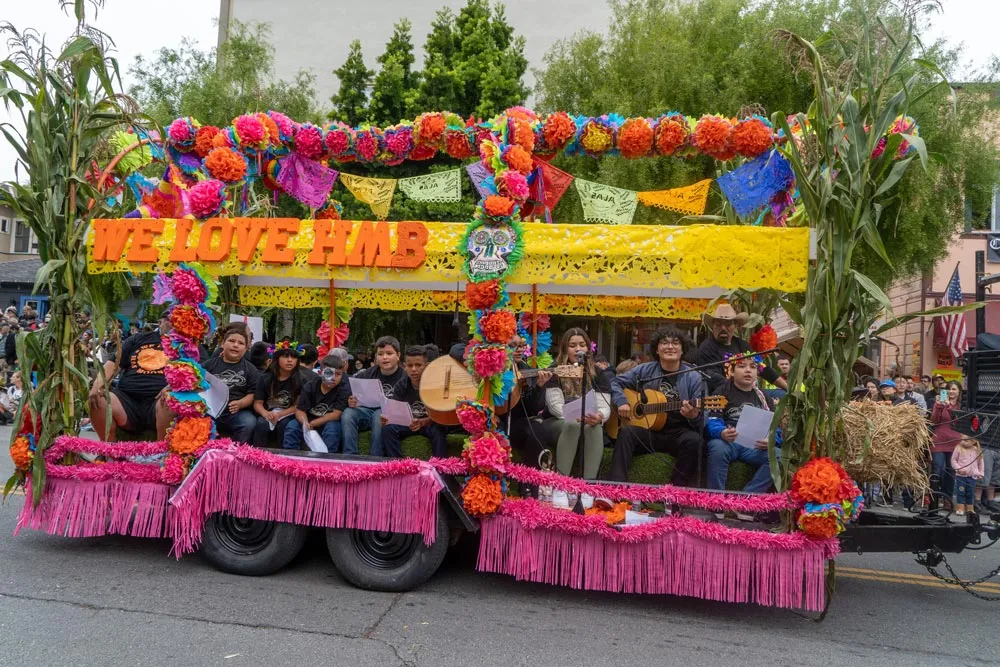 A colorful parade float decorated with flowers and corn stalks, featuring a group of children and adults singing and playing music, with the text 'WE LOVE HMB' prominently displayed.